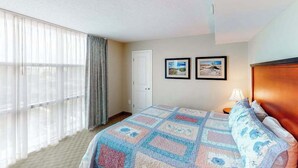 Quarto de hotel com uma cama grande, janela com cortinas leves e duas imagens emolduradas na parede.
