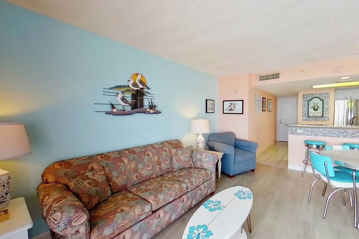Een woonkamer met een gestreept bankje, een eethoek met een tafel en stoelen, en een keuken op de achtergrond.