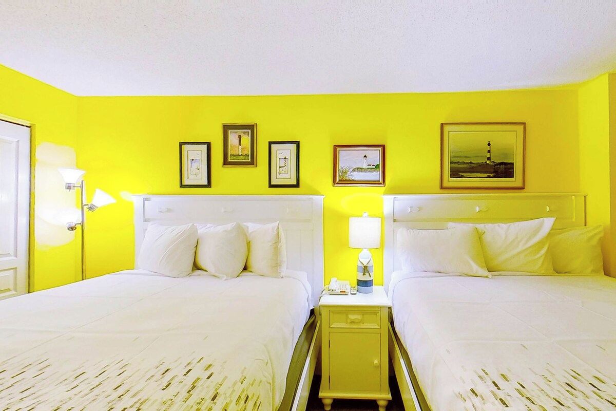 A hotel room with two beds, a nightstand, and a wall with framed pictures.