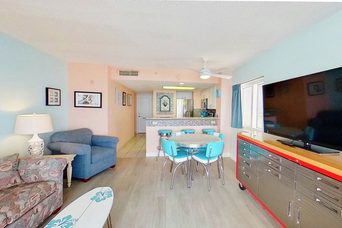 A living room with a sofa, a dining table with chairs, a television, and a kitchen area in the background.