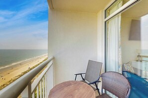 A balcony with a view of the beach, a table, two chairs, and a bed with a blue bedspread. - Bay Watch Resort 1438 (North Myrtle Beach)