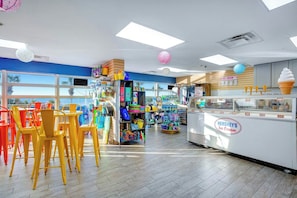 An ice cream shop with a counter displaying various flavors, a seating area with chairs, and a view of the outside through large windows. - Bay Watch Resort 1438 (North Myrtle Beach)
