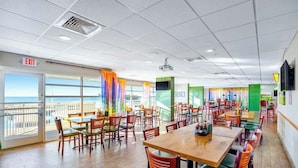 A spacious dining area with multiple tables and chairs, a view through large windows, and an exit sign. - Bay Watch Resort 0715 (North Myrtle Beach)