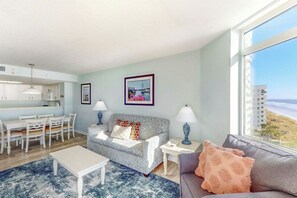 A modern living room with a sofa, a dining table, and chairs. There is a kitchen area with cabinets and a window with a view of the beach. - Bay Watch Resort 0715 (North Myrtle Beach)