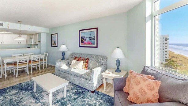 A modern living room with a sofa, a dining table, and chairs. There is a large window with a view of the beach. - Bay Watch Resort 0715 (North Myrtle Beach)
