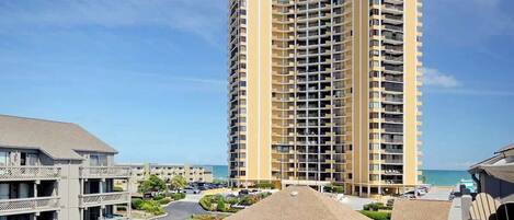 A high-rise hotel with multiple balconies, a parking lot, and a view of the beach.