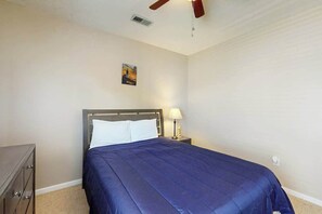 Ein Schlafzimmer mit einem Bett, einem Nachttisch mit Lampe, einem Deckenventilator und einem kleinen Gemälde an der Wand.