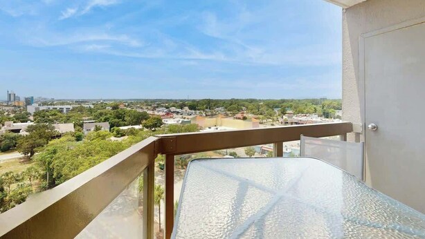 A balcony with a glass table and metal railing overlooking a cityscape with buildings and greenery. - Sea and Sails Retreat (Myrtle Beach)