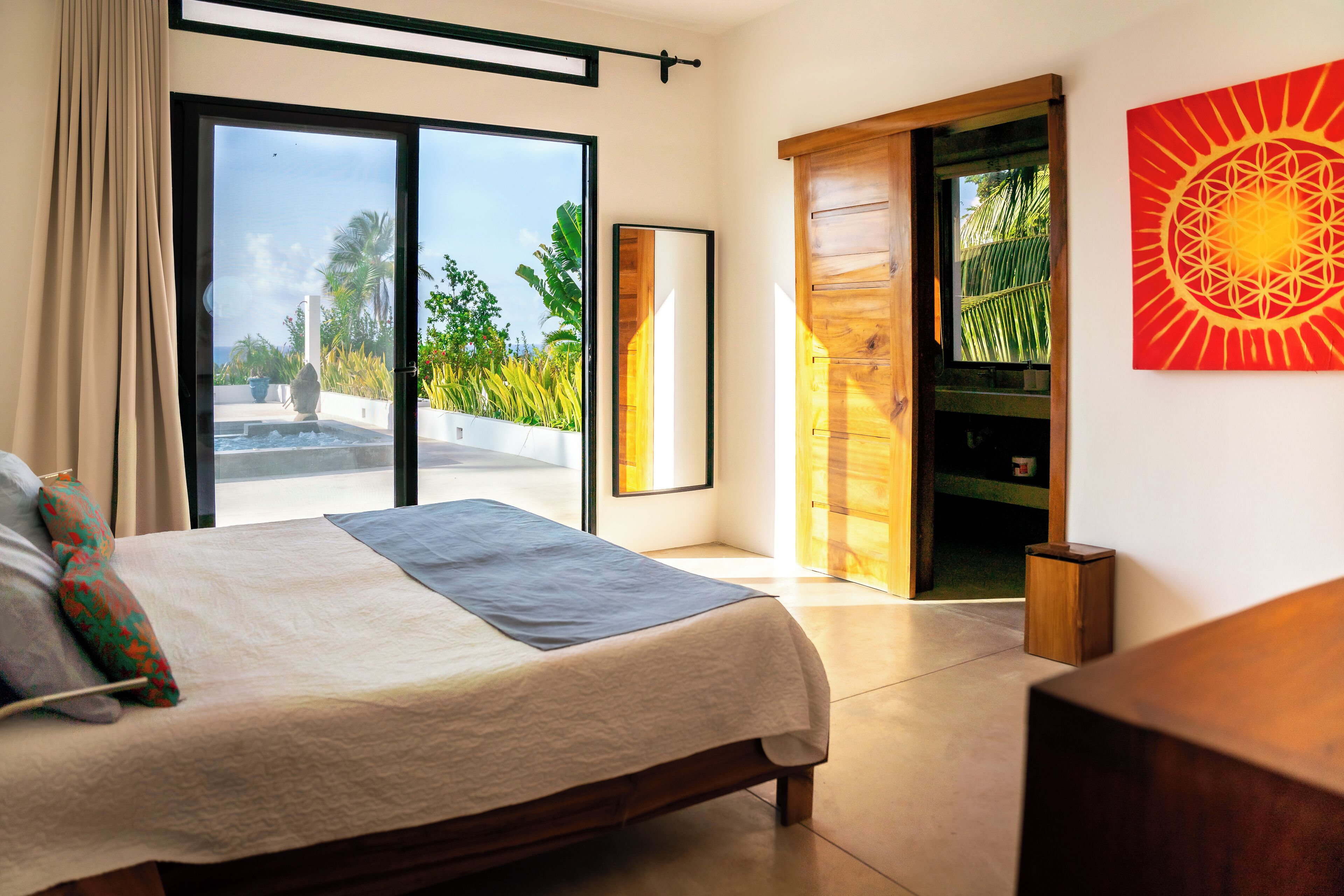 A modern bedroom with a large bed, a sliding glass door leading to a balcony, and a wooden door to a bathroom.
