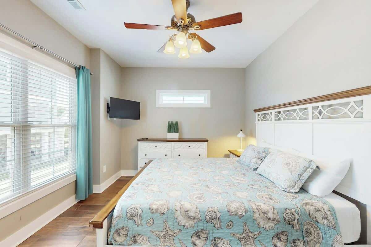 Une chambre à coucher avec un lit, un ventilateur de plafond, un téléviseur fixé au mur et une fenêtre avec des rideaux.