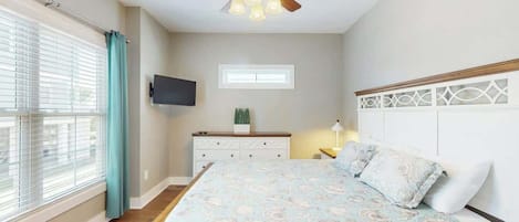 Un dormitorio con cama, ventilador de techo, televisor montado en la pared y ventana con cortinas.