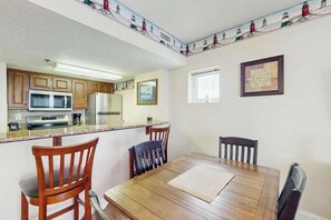 Cozinha com armários de madeira, micro-ondas e geladeira. Há uma mesa de jantar com cadeiras e uma janela com vista para faróis.