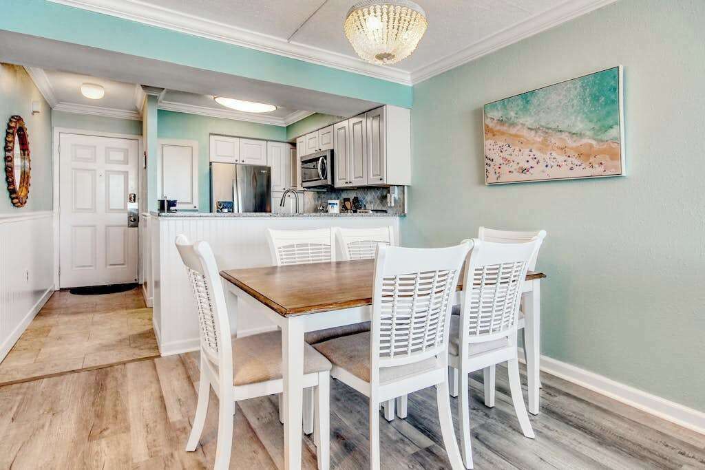 Un coin repas avec une table en bois et des chaises blanches, une cuisine avec des meubles blancs et des appareils électroménagers, et un tableau sur le thème de la plage accroché au mur.