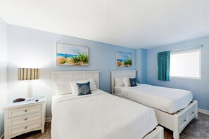 Quarto com duas camas, mesa de cabeceira, luminária e dois quadros com tema praiano.