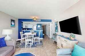 A modern living room with a sofa, a dining area with chairs, a kitchen in the background, and a flat-screen TV mounted on the wall. - Bay Watch Resort 1712 (North Myrtle Beach)