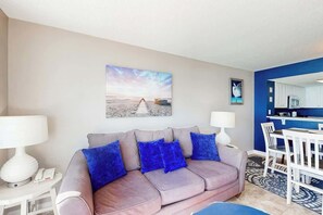 A living room with a sofa, two lamps, a painting, and a dining area with a table and chairs. - Bay Watch Resort 1712 (North Myrtle Beach)
