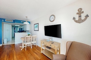 A living room with a flat-screen TV, a wooden coffee table, a brown patterned sofa, a dining area with white chairs, and a kitchen in the background. - Bay Watch Resort 1520 (North Myrtle Beach)