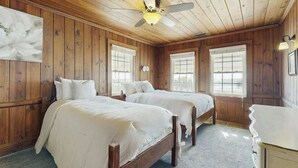 Une pièce aux murs lambrissés, avec deux lits recouverts de draps blancs, un ventilateur de plafond et une grande fenêtre.
