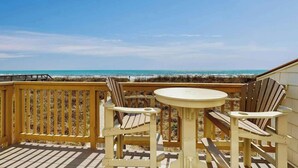 Une terrasse en bois avec une table ronde et deux chaises Adirondack donnant sur une plage.