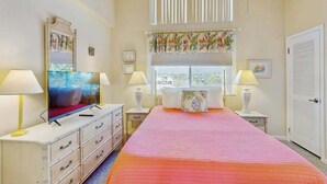 Une chambre à coucher comprenant un lit, deux tables de chevet, un téléviseur et une fenêtre avec des stores.
