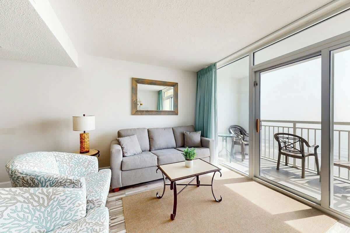 Een moderne woonkamer met een bank, fauteuil en salontafel, een balkon met stoelen en een groot raam.