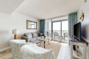 A living room with a sofa, two armchairs, a coffee table, a TV, and a balcony with chairs. - Camelot by the Sea 0905 (Myrtle Beach)