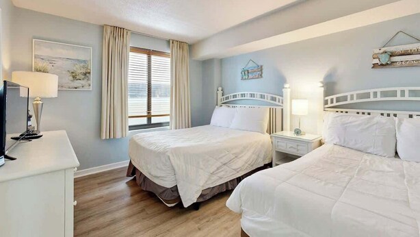 A hotel room with two beds, a desk, a lamp, a window with curtains, and a wall-mounted decoration. - Golden Mile Condo Retreat (Myrtle Beach)