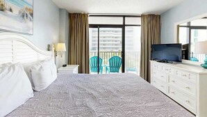 A hotel room with a bed, a television, a dresser, a balcony with chairs, and a painting on the wall. - Golden Mile Condo Retreat (Myrtle Beach)