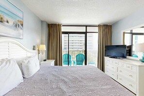 Ein Hotelzimmer mit Bett, Fernseher, Kommode, einem Balkon mit Stühlen und einem Bild an der Wand.
