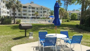 Eine Terrasse im Freien mit einem runden Tisch, blauen Stühlen, einem Grill und einem Pool im Hintergrund.