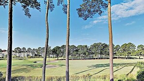 Ein Golfplatz mit Bäumen, blauem Himmel und Gebäuden im Hintergrund.