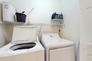 A laundry area with a front-loading washer and dryer, a shelf with cleaning supplies, and a bucket. - Magnolia Pointe Unit 204 (Myrtle Beach)