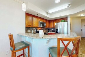 A kitchen with wooden cabinets, a granite countertop, a stainless steel refrigerator, and two wooden chairs with green cushions. - The Horizon at 77th Unit 805 (Myrtle Beach)