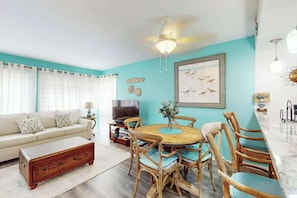 A living room with a sofa, coffee table, dining table with chairs, and a TV. - Arcadian Dunes 12-246 (Myrtle Beach)