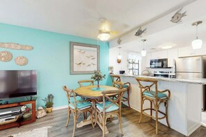 A modern kitchen with white cabinets, a stainless steel refrigerator, and a dining area with wooden chairs and a round table. - Arcadian Dunes 12-246 (Myrtle Beach)