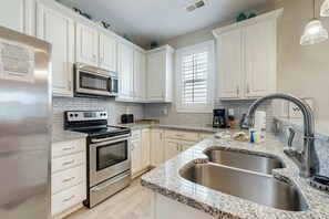 A kitchen with white cabinets, a stainless steel refrigerator, a double oven, a microwave, a granite countertop, a double sink, and a pendant light. - The Retreat Condo Home (North Myrtle Beach)