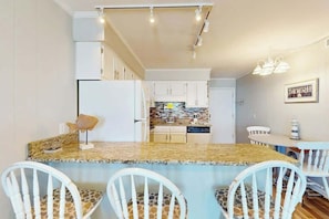 A kitchen with white cabinets, a granite countertop, and a dining area with white chairs. - A Place at the Beach VI - 305 (Myrtle Beach)