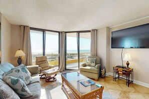 A hotel room with a large window, a flat-screen TV, a sofa, a chair, a small table, and a lamp. - Ocean Creek Plantation D-5 (Myrtle Beach)