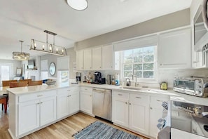 A modern kitchen with white cabinetry, a built-in dishwasher, a stainless steel oven, and a window providing natural light. - Coastal Gem of Murrells Inlet (Murrells Inlet)