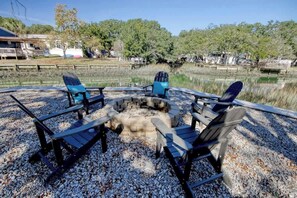 A circular seating arrangement with a fire pit in the center, surrounded by chairs, set on a gravel surface with a body of water and trees in the background. - Coastal Gem of Murrells Inlet (Murrells Inlet)