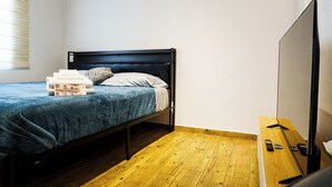 A single bed with a dark headboard, a blue blanket, and a white pillow. There is a wooden nightstand with a flat-screen TV and remote controls. The room has wooden flooring and a window with blinds. - Matlazinca Suite in downtown Toluca (Toluca de Lerdo)
