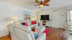 A living room with a sofa, television, ceiling fan, and a door. - Shorehaven G-3 Unit Condo (North Myrtle Beach)