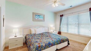 Un dormitorio con cama, mesitas de noche, ventilador de techo, ventana con cortinas e imagen enmarcadas en la pared.