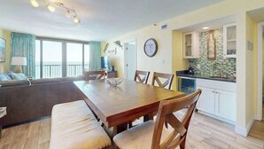 A dining area with a wooden table and chairs, a sofa, and a kitchen with a tiled backsplash. - Sea Winds PH4 Unit Condo (North Myrtle Beach)