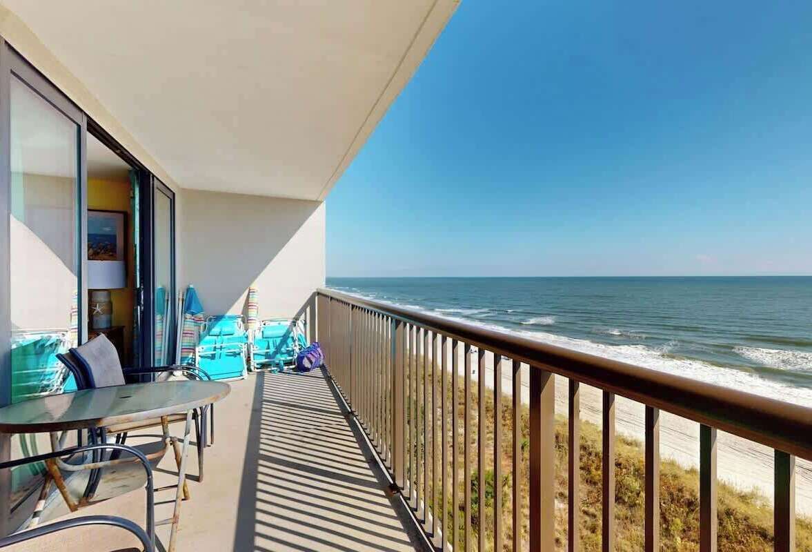 A balcony with a table and chairs, overlooking a beach and the ocean.