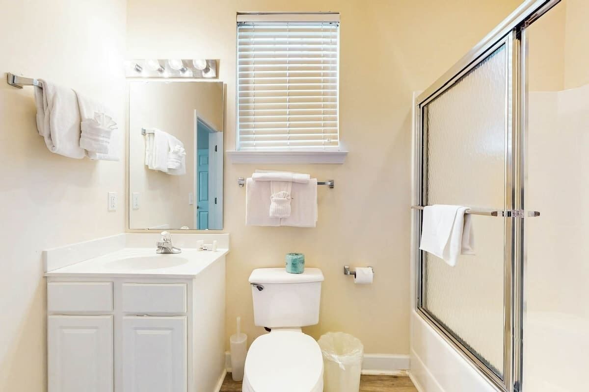 Un baño con lavamanos, inodoro y ducha blancos. Hay toallas colgadas en la pared y una ventana con persianas.