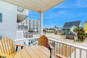 A balcony with wooden chairs and a table, overlooking a street with buildings and palm trees. - Seaside Unit C Condo (Surfside Beach)