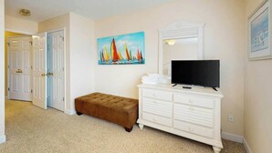 Una habitación con un televisor sobre un tocador blanco, un cuadro de veleros en la pared y un banco marrón.