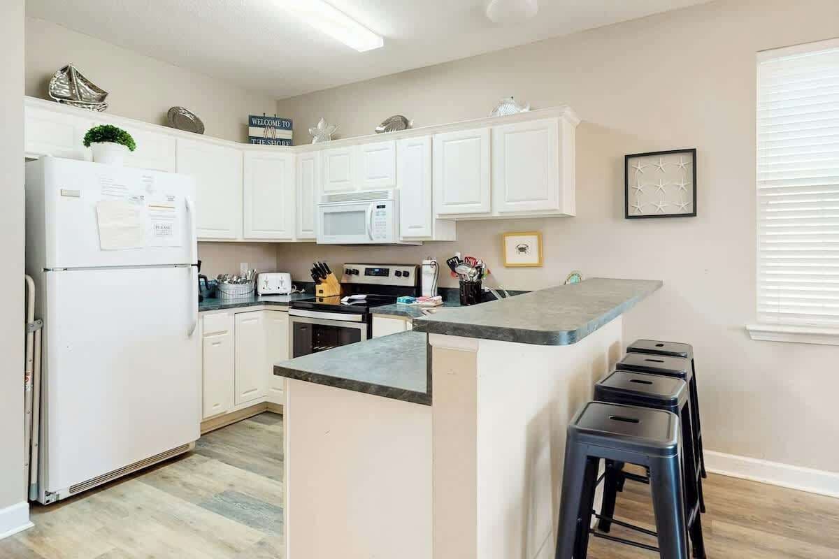 A modern kitchen with white cabinets, a built-in microwave, a stove, and a bar area with stools.