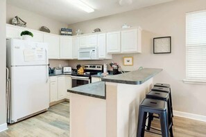 A modern kitchen with white cabinets, a built-in microwave, a stove, and a bar area with stools. - Seaside Unit C Condo (Surfside Beach)